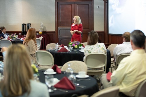 female speaker presenting to crowd of people seated at tables